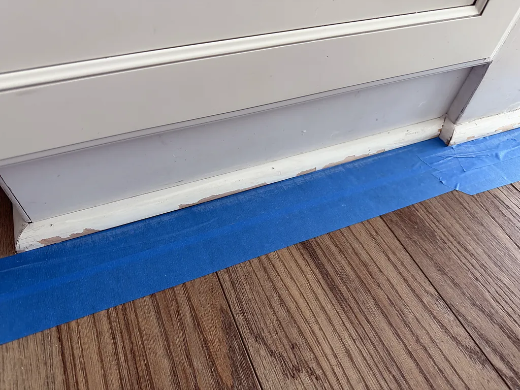 Baseboard masked with blue painter's tape against wood flooring before repainting