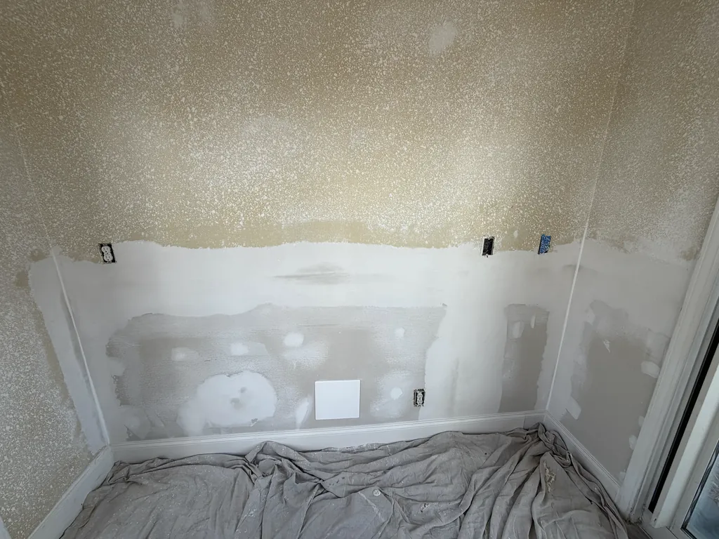 Textured wall partway through repair, with fresh patches of joint compound along the lower half and drop cloths on the floor